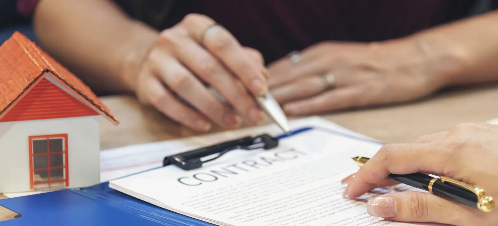 Hands signing a contract on a clipboard next to a small model house